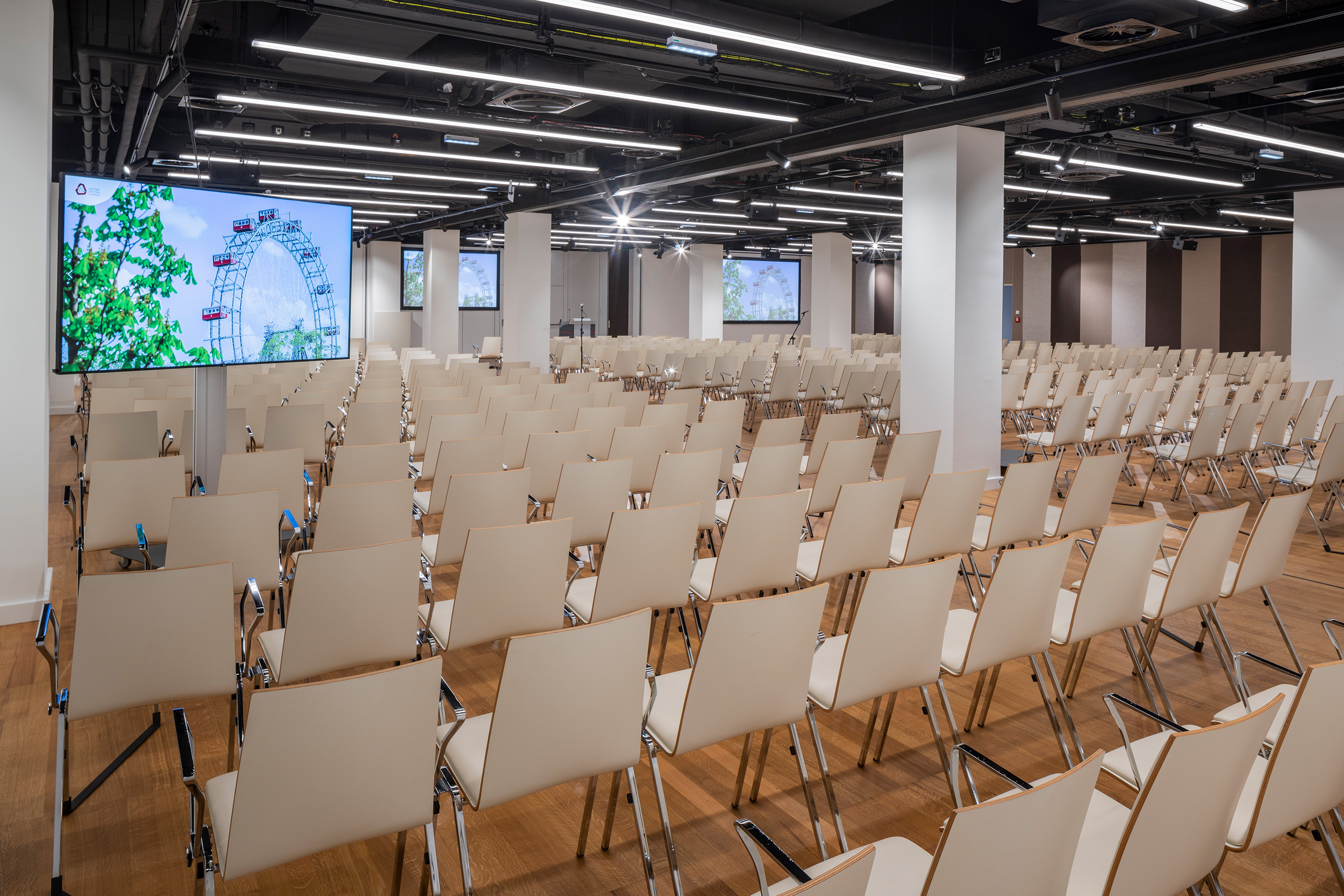 Foto: Sitzreihen in einem Saal mit Holzboden, Monitore, gestreifte Tapete