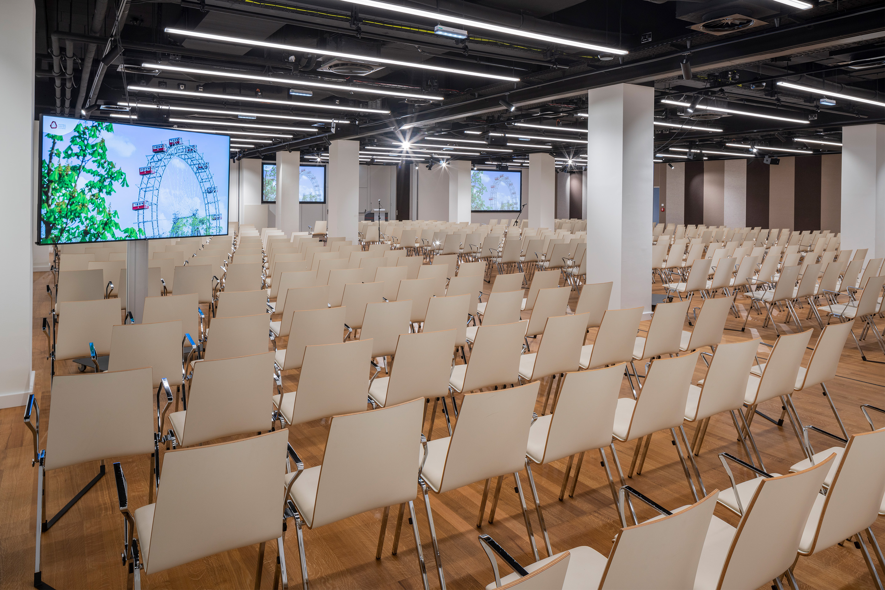 Foto: Sitzreihen in einem Saal mit Holzboden, Monitore, gestreifte Tapete