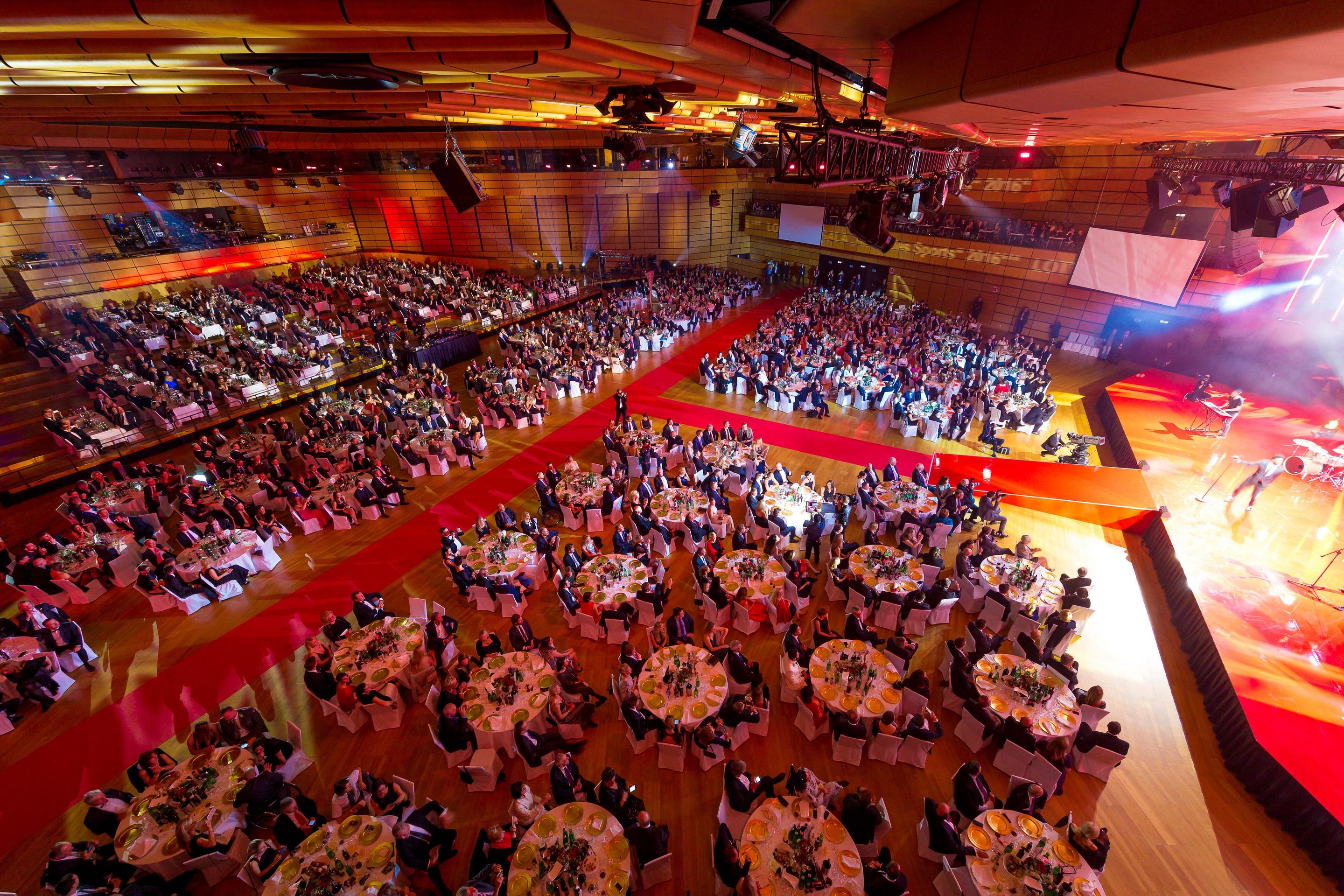 Foto: Menschen in einer Halle auf Gala-Tischen, roter Teppich, Buehne