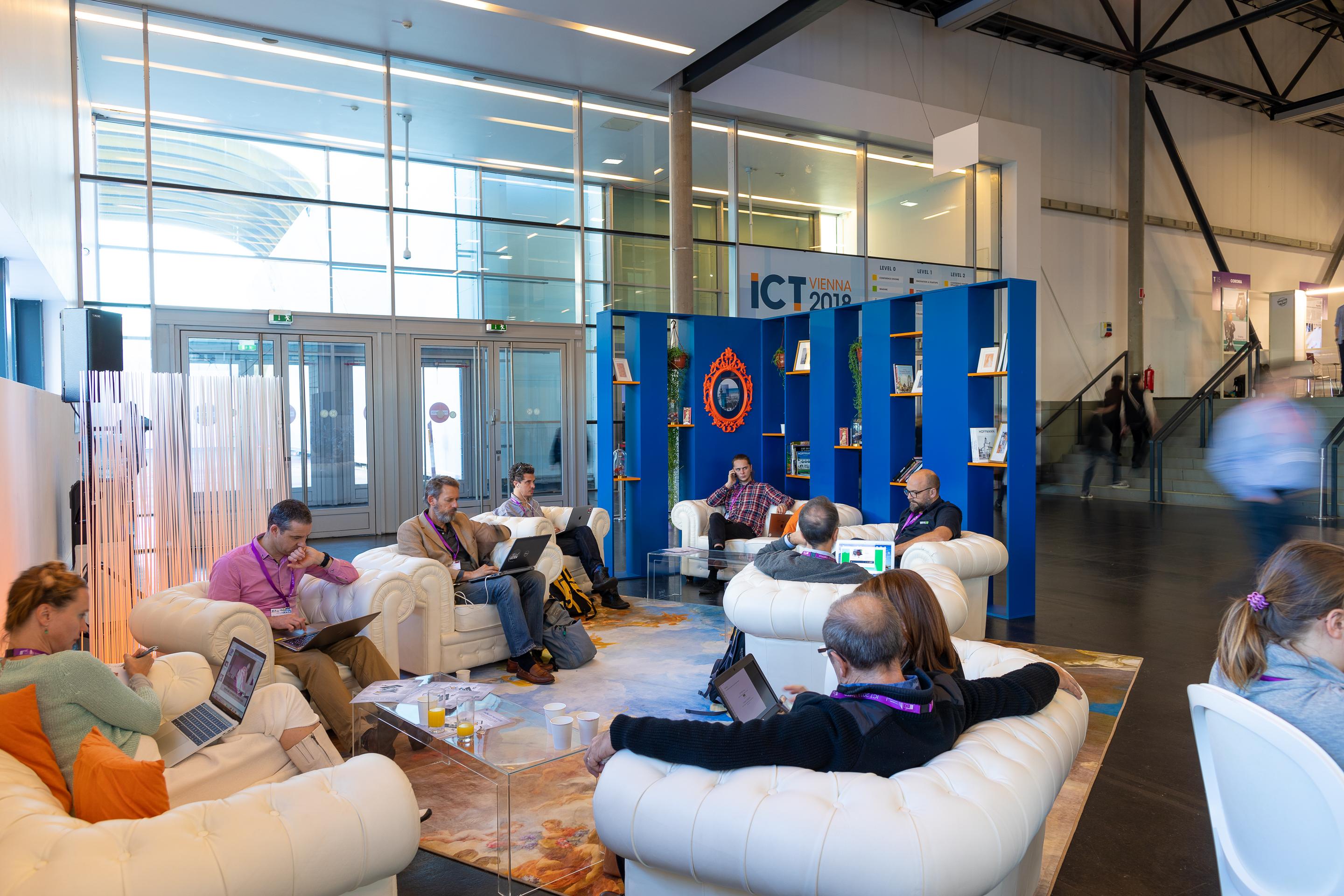 Foto: Menschen in Sitzgruppen bei einem blauen Buchregal in einer Halle