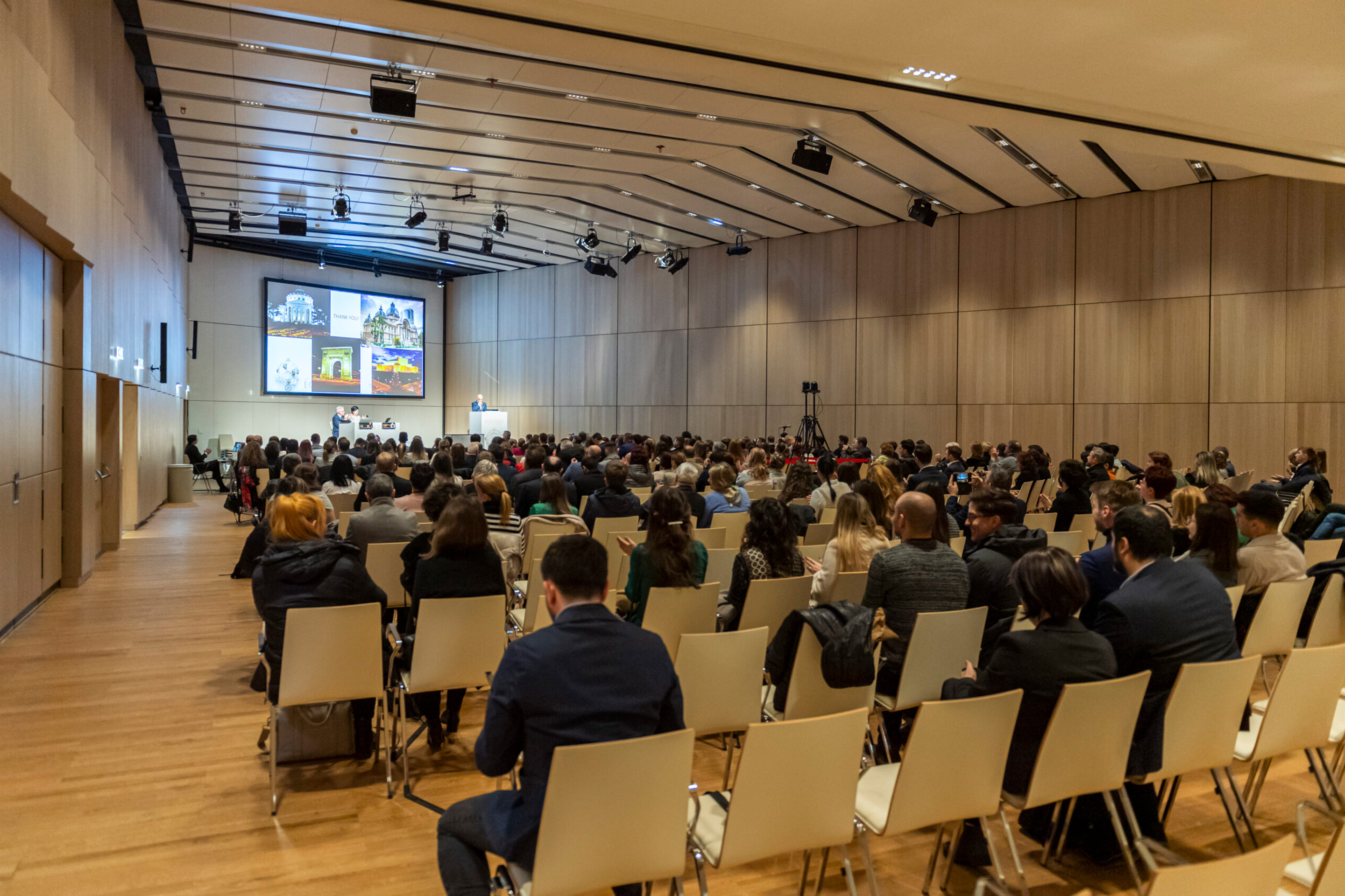 Foto: Holzener Saal voller Stuehle und Menschen bei einem Vortrag