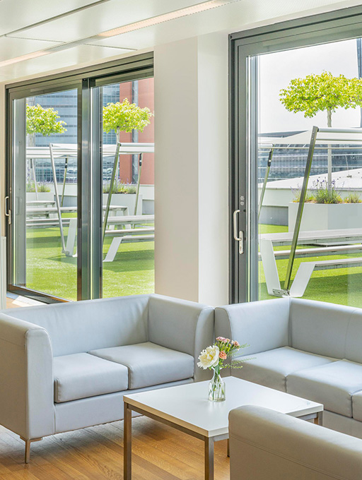 Foto: Lounge Moebel mit Balkontueren. Im Hintergrund ist eine Terrasse mit Sitzbaenken, Baeumen und gruenem Teppich.