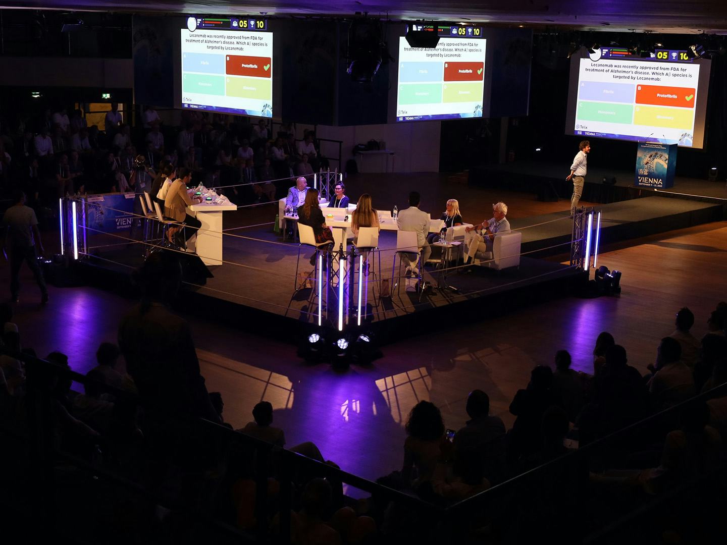 Foto: dunkler Raum mit Publikum und beleuchtetem Podium mit Diskussionsteilnehmern und Praesentation auf Leinwaenden