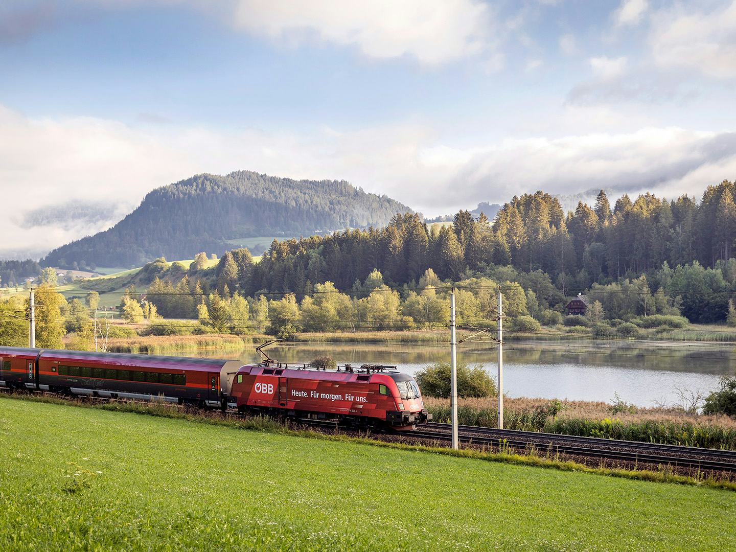 Bild: roter Zug faehrt durch Landschaft, im Hintergrund ein See und Wald
