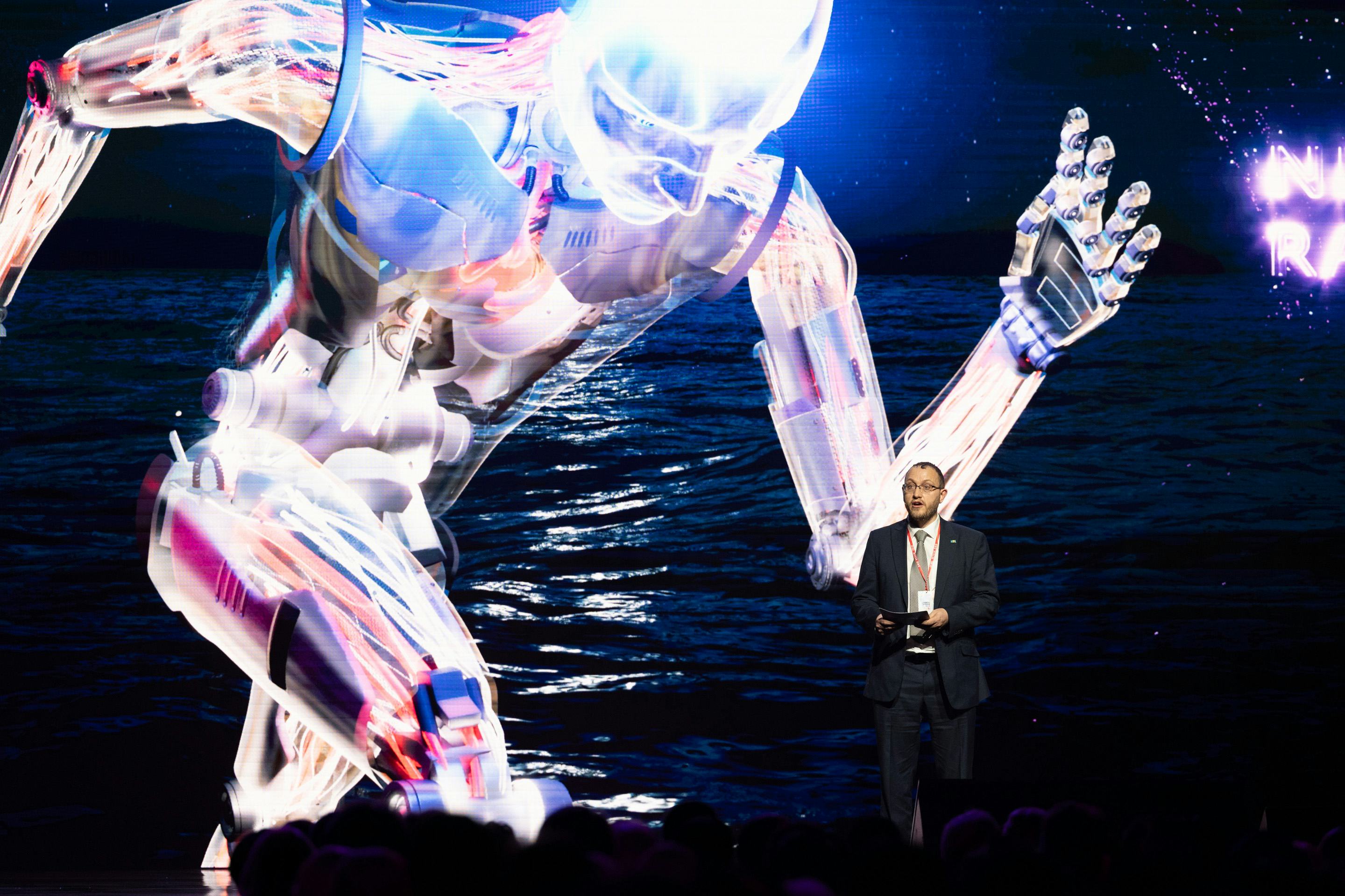 Foto: eine Leinwand, auf der ein laufender Roboter zu sehen ist. Im Vordergrund ein Moderator.