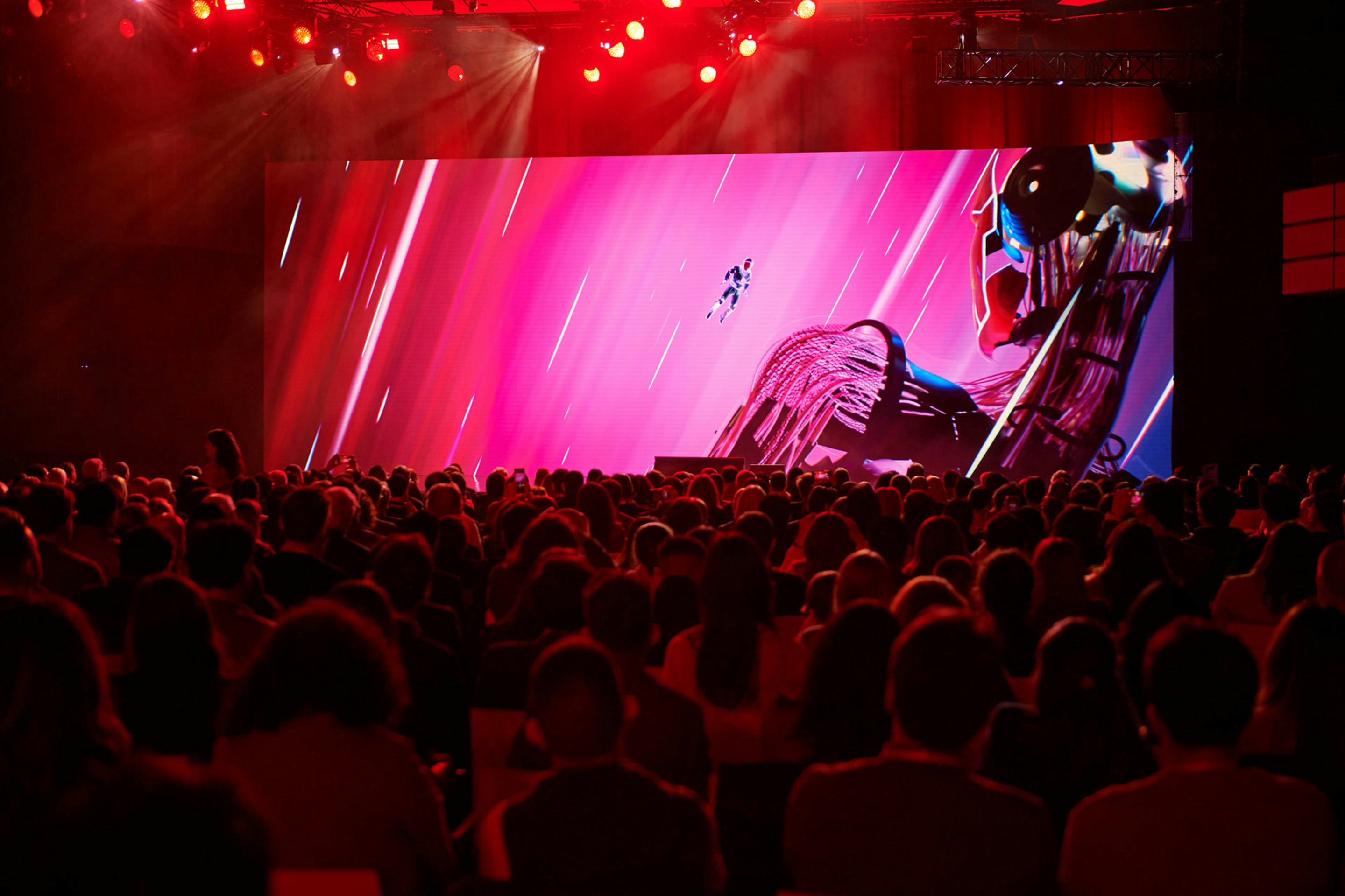 Foto: rot beleuchteter Saal voller Menschen, im Hintergrund eine Buehne und eine Show auf einer LED Waende