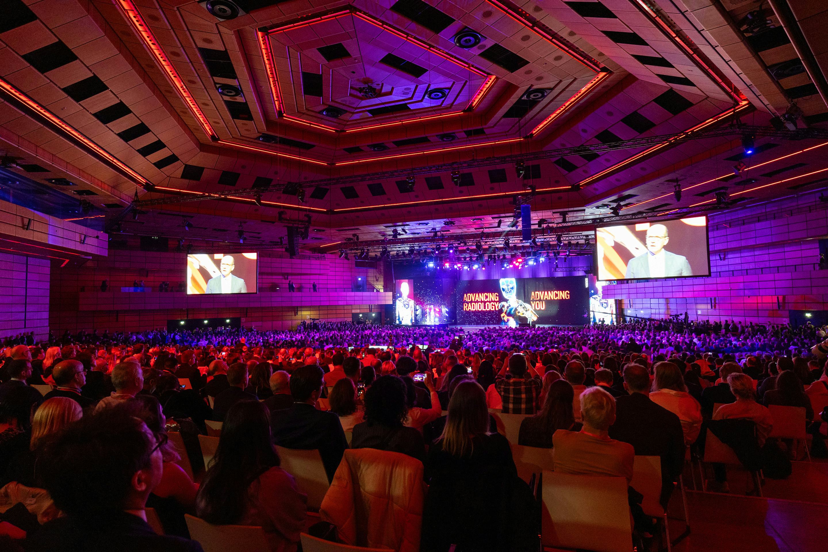 Foto: rot beleuchteter Saal voll sitzender Menschen, die einem Moderator auf der Buehne zuhoeren.