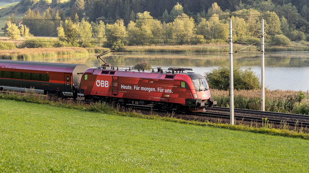Roter OEBB Zug faehrt durch Landschaft, im Hintergrund ein See und Wald.