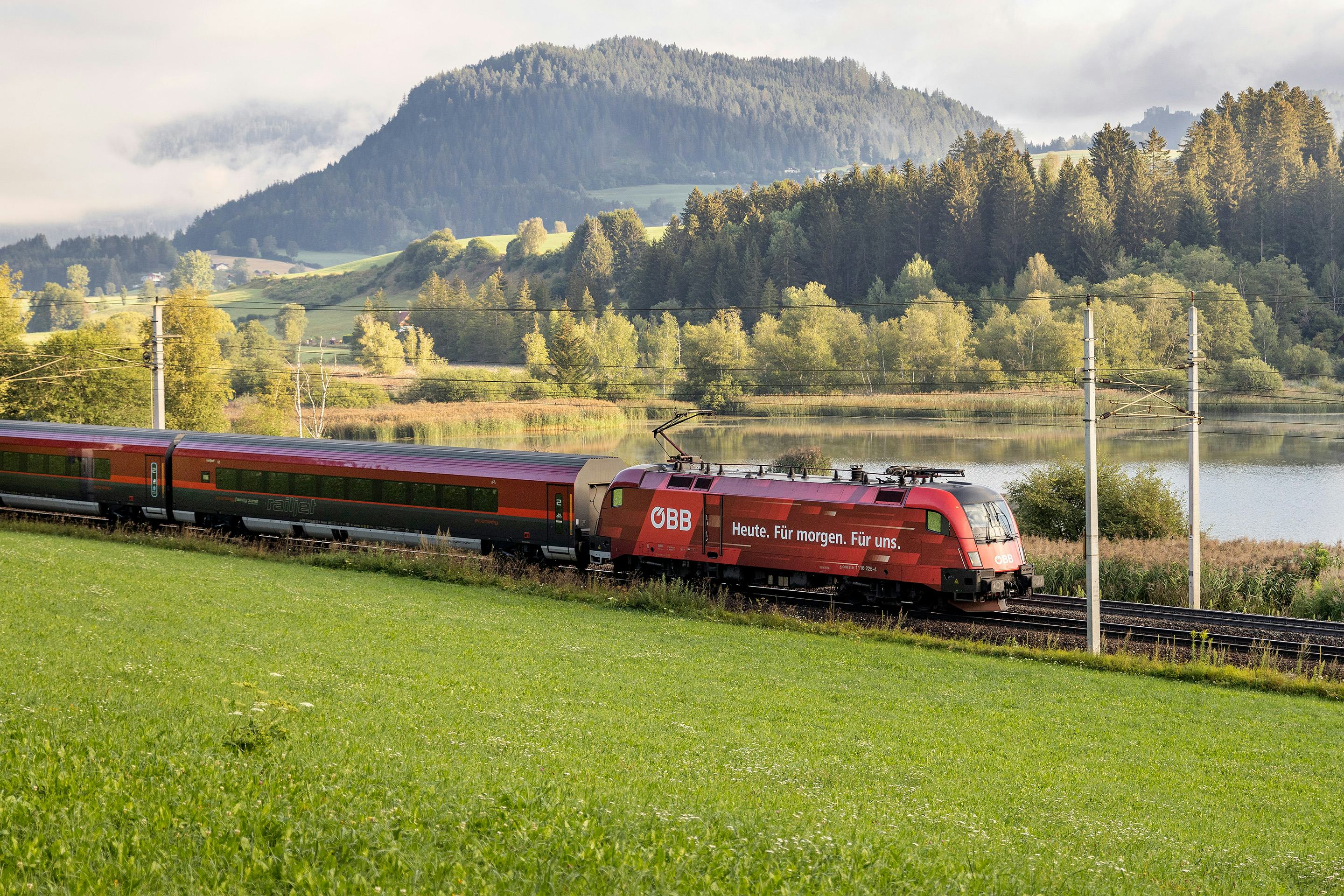 Bild: roter Zug faehrt durch Landschaft, im Hintergrund ein See und Wald