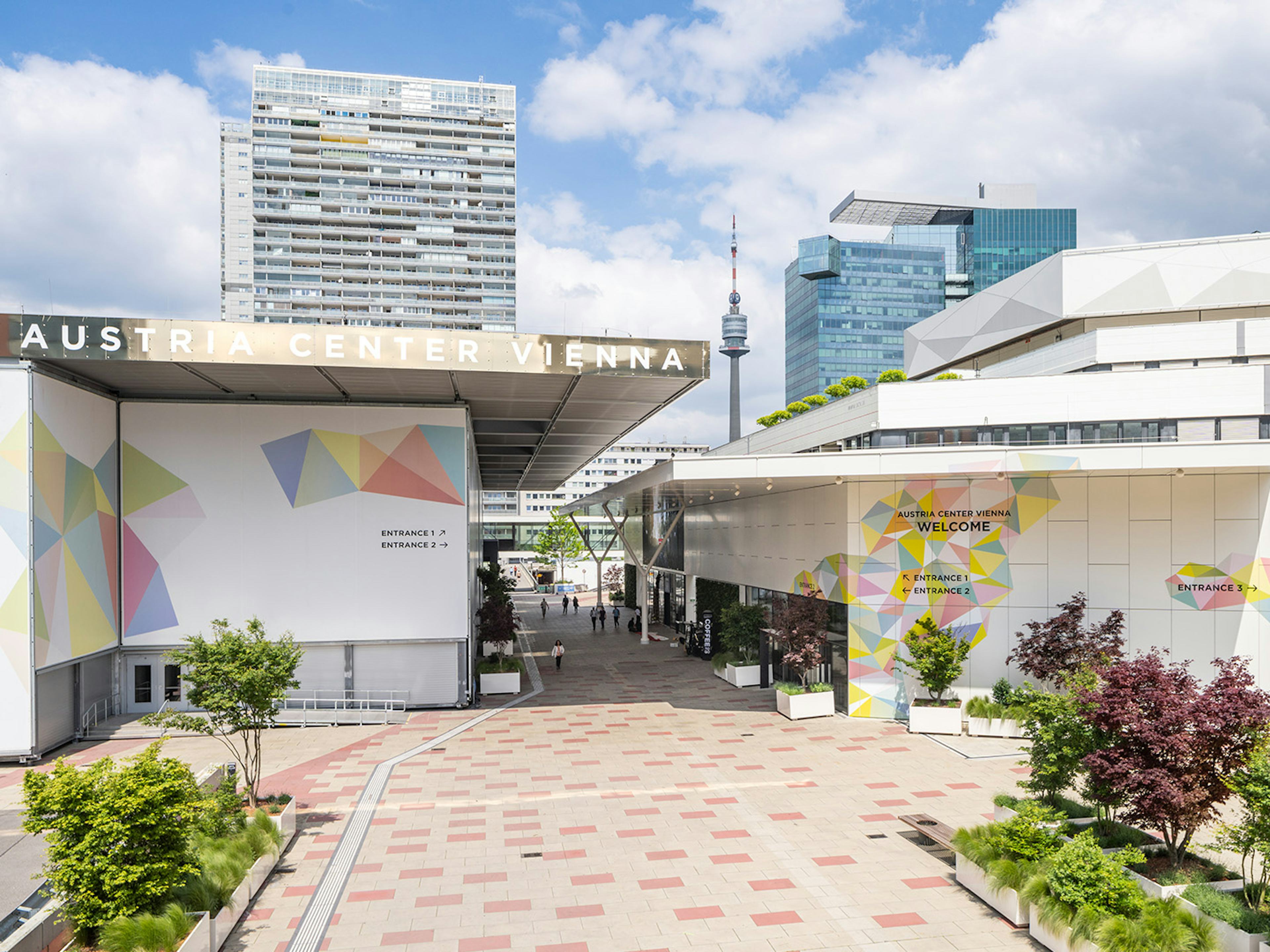 Aussenansicht des Austria Center Vienna mit Hochhaeusern und Donauturm im Hintergrund.