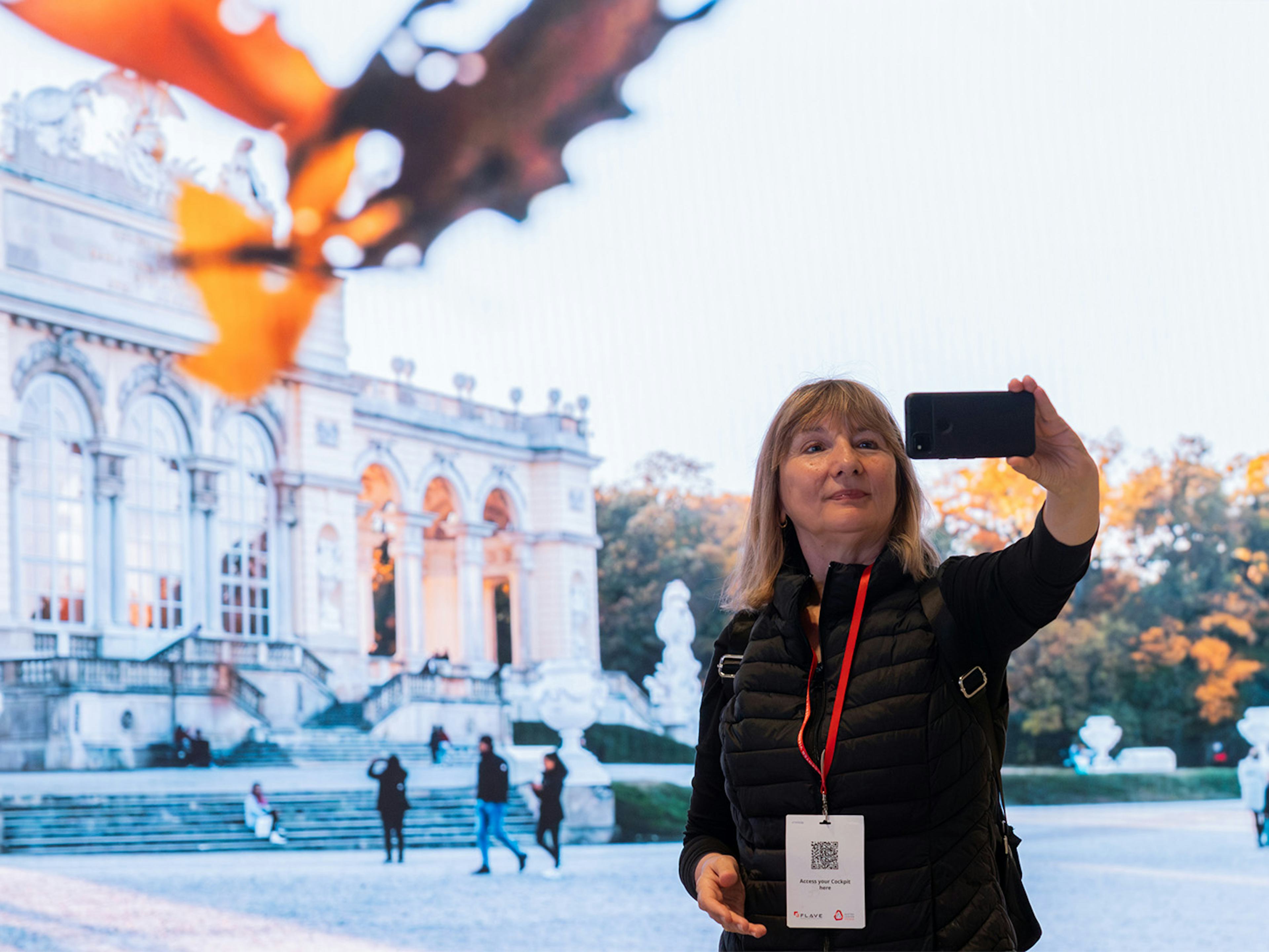 Frau posiert fuer Selfie vor einer LED-Wand, auf der die Gloriette in Park Schoenbrunn zu sehen ist.