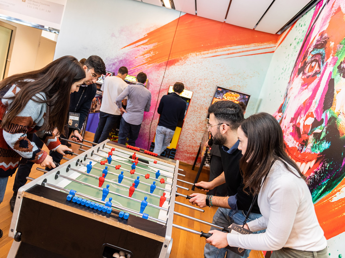 Zwei Frauen und zwei Maenner spielen Tischfussball, im Hintergrund stehen Maenner an Spieleautomaten.